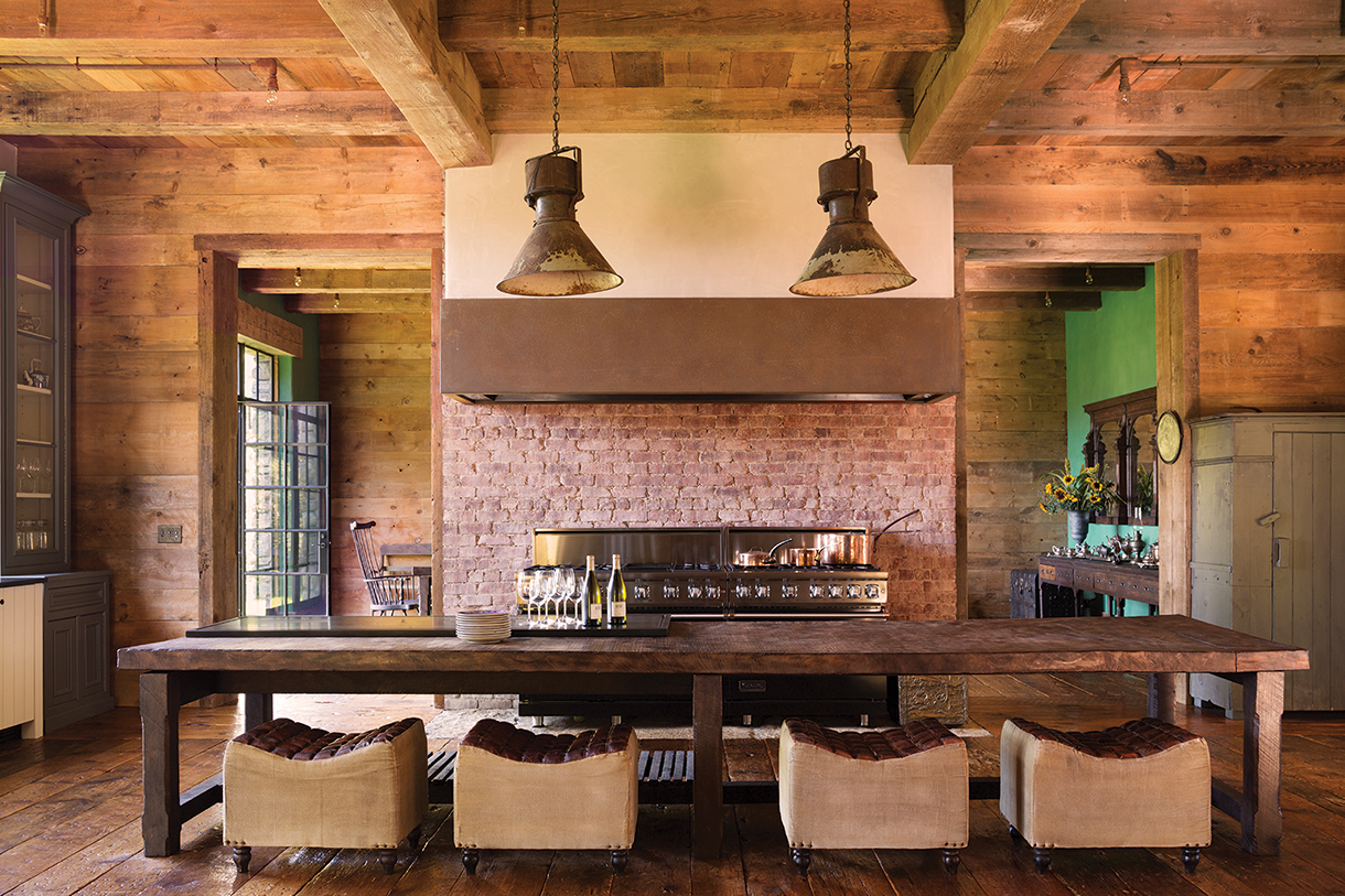 English manor house kitchen with reclaimed brick fireplace, wood flooring, worktable island, and antique chairs.